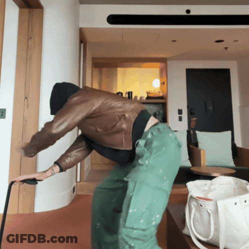 A Man In A Leather Jacket And Green Pants Is Standing In A Room GIF