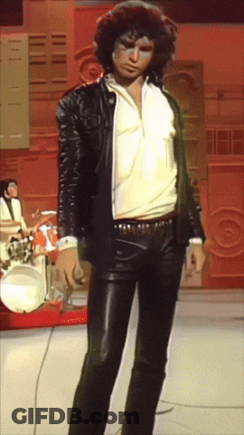 A Man In A Leather Jacket And Pants Stands In Front Of Drums GIF