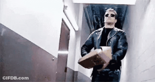 A Man In A Leather Jacket And Sunglasses Is Carrying A Cardboard Box In A Hallway . GIF