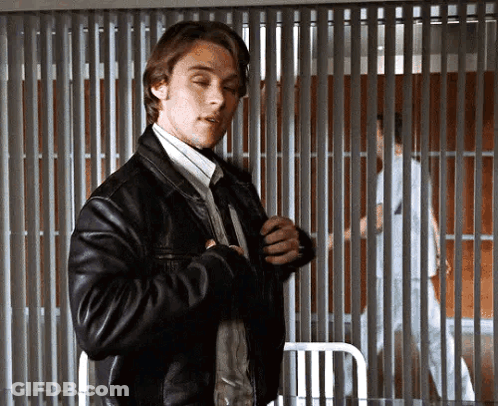 A Man In A Leather Jacket And Tie Is Standing In Front Of A Prison Cell . GIF