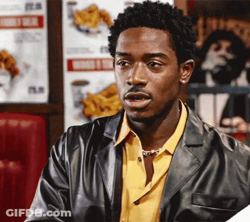 A Man In A Leather Jacket And Yellow Shirt Is Sitting In A Diner GIF
