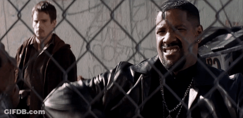 A Man In A Leather Jacket Is Behind A Chain Link Fence With Another Man Behind Him GIF