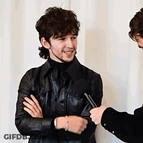 A Man In A Leather Jacket Is Being Interviewed By A Microphone While Standing Next To Another Man . GIF