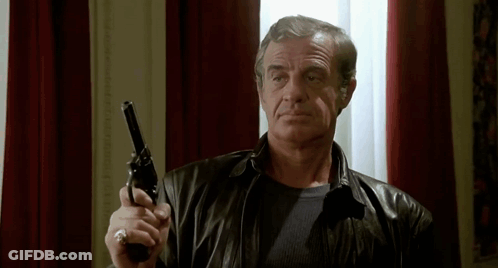 A Man In A Leather Jacket Is Holding A Gun In His Right Hand . GIF