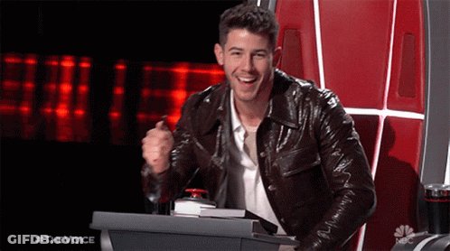 A Man In A Leather Jacket Is Sitting At A Podium With A Red Button That Says #Thevoice GIF