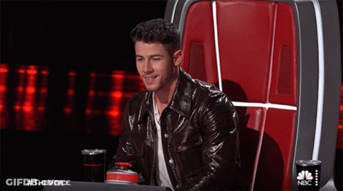 A Man In A Leather Jacket Is Sitting In A Chair With A Nbc Logo On The Back GIF
