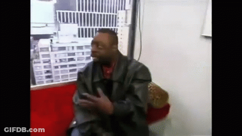 A Man In A Leather Jacket Is Sitting On A Red Couch And Talking On A Cell Phone . GIF
