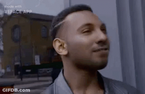 A Man In A Leather Jacket Is Smiling And Looking At The Camera While Standing In Front Of A Building . GIF