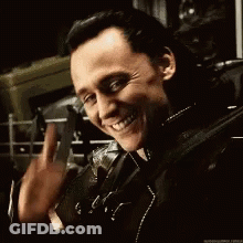 A Man In A Leather Jacket Is Smiling And Waving While Holding A Gun . GIF