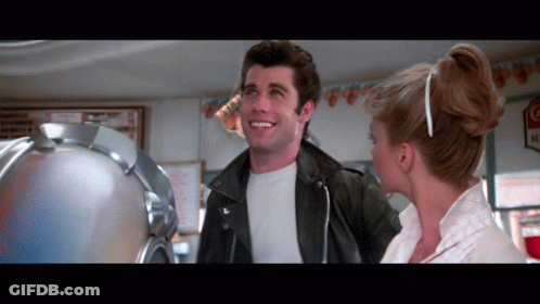 A Man In A Leather Jacket Is Smiling Next To A Woman In A White Dress GIF