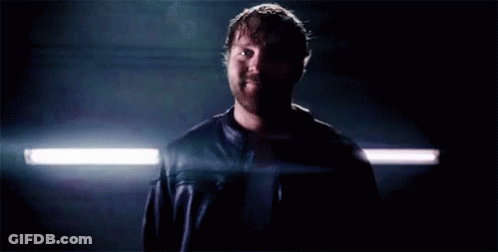 A Man In A Leather Jacket Is Standing In A Dark Room With A Light Shining On Him . GIF