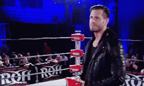 A Man In A Leather Jacket Is Standing In A Wrestling Ring With A Sign That Says Rqf GIF