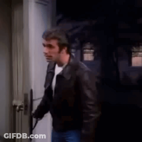 A Man In A Leather Jacket Is Standing In Front Of A Door And Holding A Gun . GIF