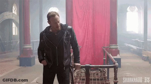 A Man In A Leather Jacket Is Standing Next To A Walker In A Room With A Red Curtain Behind Him . GIF