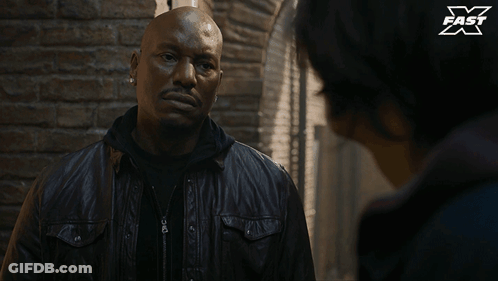 A Man In A Leather Jacket Is Talking To Another Man In Front Of A Brick Wall With X Fast Written On It GIF