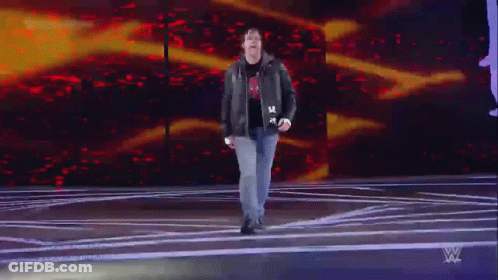 A Man In A Leather Jacket Is Walking On A Stage In Front Of A WWE Logo GIF