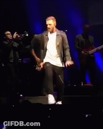 A Man In A Leather Jacket Is Walking On A Stage With A Woman Behind Him GIF