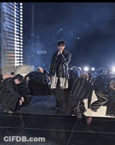 A Man In A Leather Jacket Sings Into A Microphone In Front Of A Crowd GIF