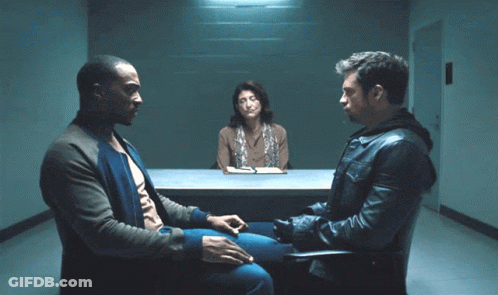 A Man In A Leather Jacket Sits In A Chair Talking To Two Other Men GIF