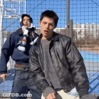 A Man In A Leather Jacket Stands Next To Another Man In A Scarf GIF