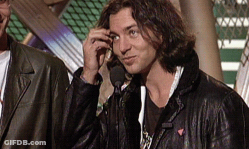 A Man In A Leather Jacket Talks Into A Microphone While Another Man Stands Behind Him GIF