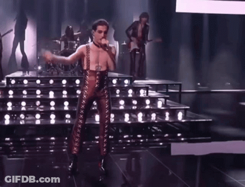 A Man In A Leather Outfit Is Singing Into A Microphone On Stage . GIF