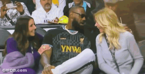 A Man In A Lebron James Jersey Is Sitting In A Stadium With Two Women . GIF