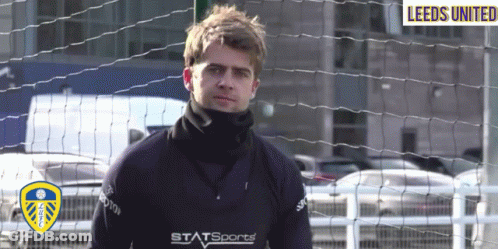 A Man In A Leeds United Jersey Stands Behind A Fence GIF