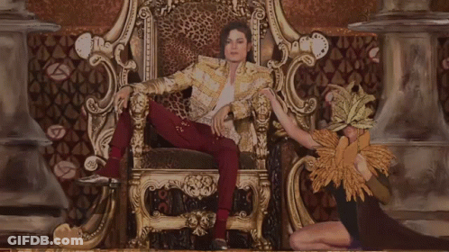 A Man In A Leopard Print Jacket Is Sitting On A Throne With A Woman Behind Him GIF