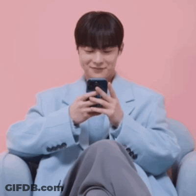 A Man In A Light Blue Coat Is Sitting In A Chair Looking At His Phone GIF