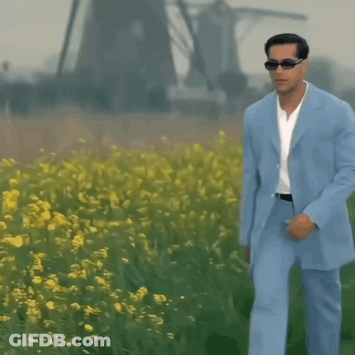 A Man In A Light Blue Suit And Sunglasses Walks Through A Field Of Yellow Flowers GIF
