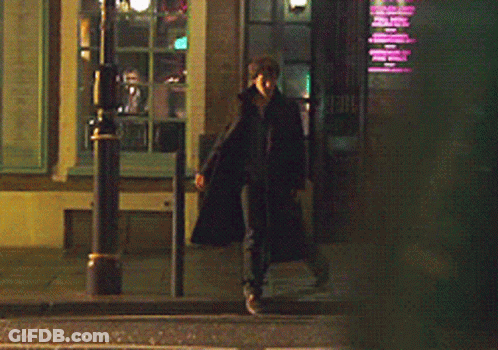 A Man In A Long Black Coat Is Walking Down A Street At Night GIF
