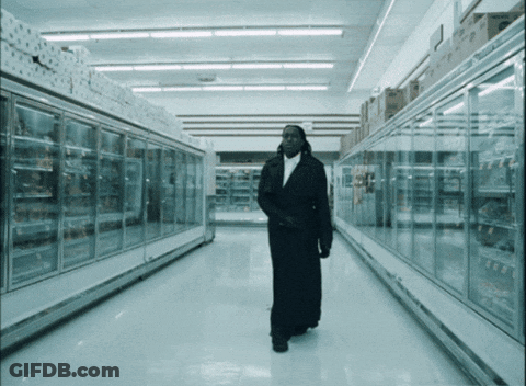 A Man In A Long Black Coat Walks Through An Empty Supermarket Aisle GIF
