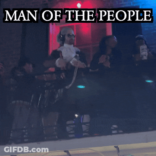 A Man In A Man Of The People Costume Stands In Front Of A Green Light GIF
