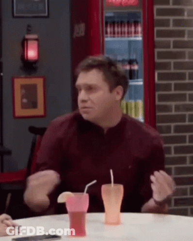 A Man In A Maroon Shirt Is Sitting At A Table With Two Drinks In Front Of Him . GIF
