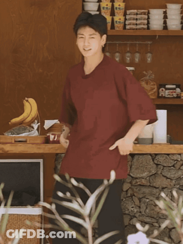 A Man In A Maroon Shirt Is Standing In Front Of A Shelf With Bowls On It GIF