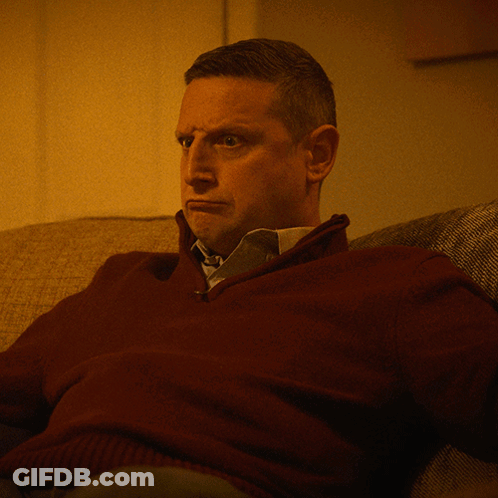 A Man In A Maroon Sweater And Tie Sits On A Couch GIF