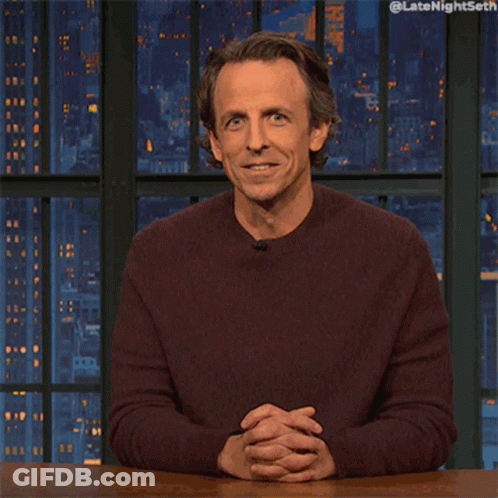 A Man In A Maroon Sweater Is Sitting At A Table With His Hand On His Chin And The Words Late Night Seth On The Bottom GIF
