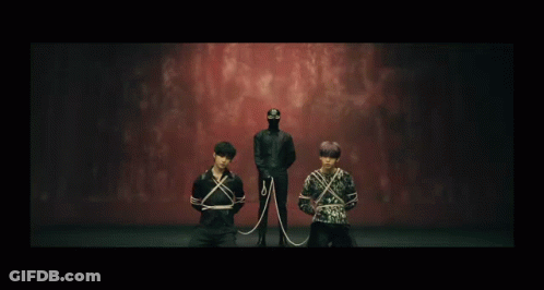 A Man In A Mask Is Standing Behind Two Men Tied Up With Ropes GIF
