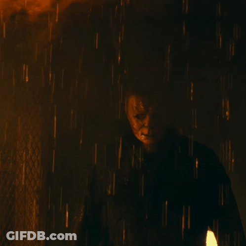 A Man In A Mask Is Standing In The Rain With A Fire In The Background GIF