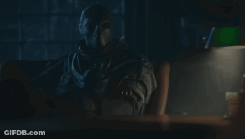 A Man In A Mask Sits At A Desk With A Lamp GIF