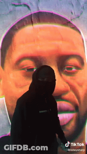 A Man In A Mask Stands In Front Of A Large Painting Of A Man 'S Face GIF