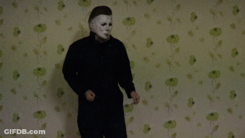 A Man In A Mask Stands In Front Of A Wall With Green Flowers On It GIF