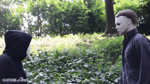 A Man In A Mask Stands Next To Another Man In A Hood GIF