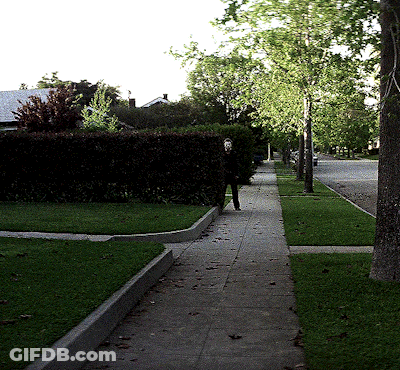 A Man In A Mask Walks Down A Sidewalk In A Neighborhood GIF