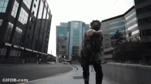 A Man In A Mask Walks Down A Street In Front Of A Building GIF
