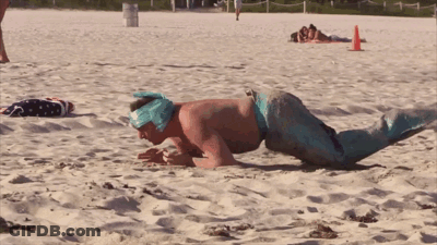 A Man In A Mermaid Costume Is Doing Push Ups On A Sandy Beach GIF