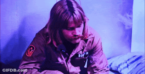 A Man In A Military Jacket With The Word Bof On His Chest Is Sitting On A Bed . GIF