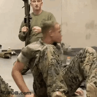 A Man In A Military Uniform Is Sitting On The Floor While Another Man Holds A Rifle . GIF