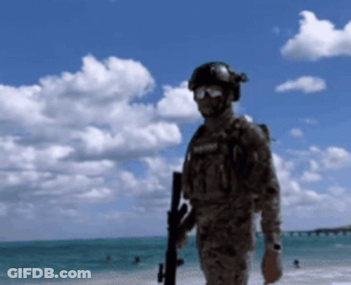 A Man In A Military Uniform Is Standing On A Beach Holding A Gun GIF